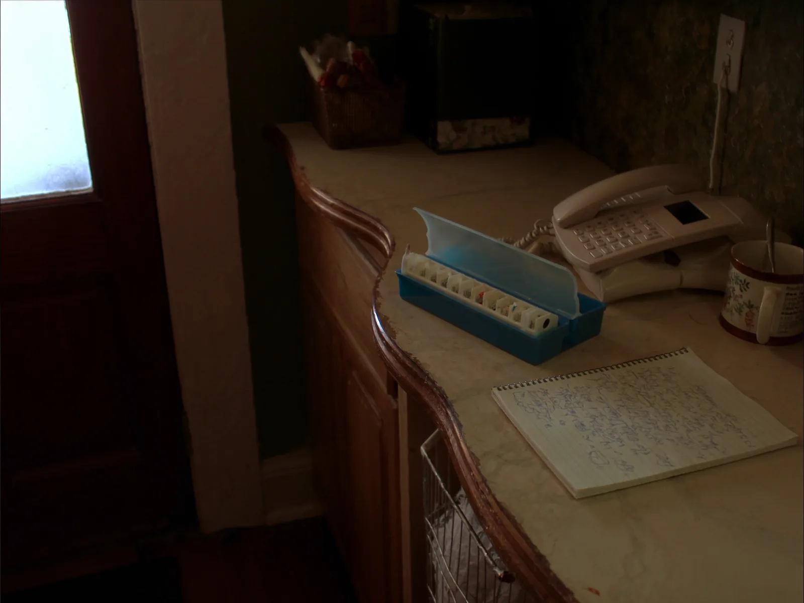 A kitchen counter in an older home with a seven day pill organiser part filled, an open spiral notebook, a landline phone and a coffee mug.