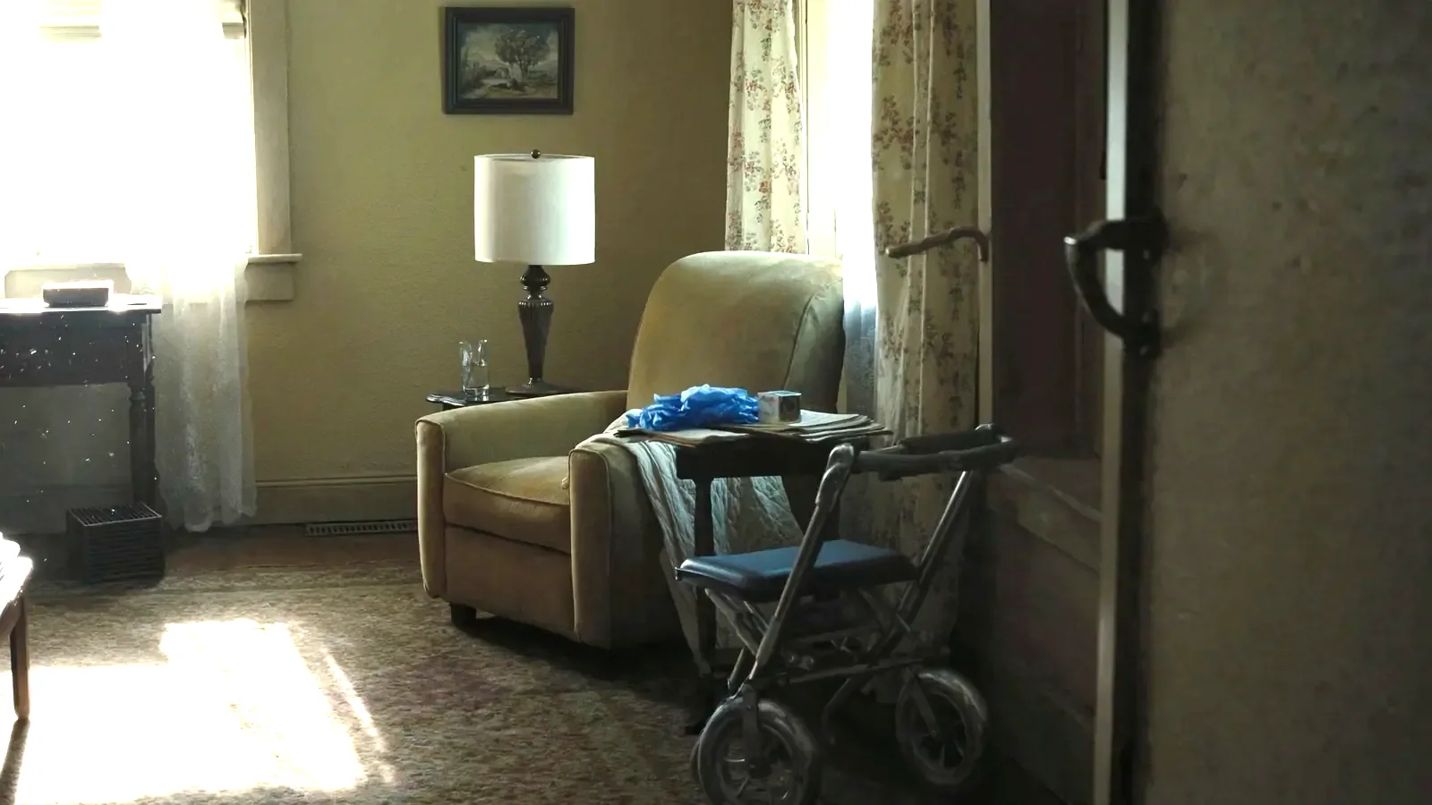A sunlit front room with an empty armchair, a folded quilt over the arm and a wheeled walker parked beside it.